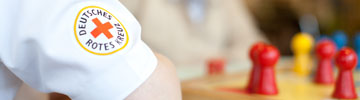 Mehr Inofmationen zum freiwilligen Engagement in unserem Ortsverein. Foto: Arm einer DRK-Pflegerin vor einem Mensch-Ärger-dich-nicht-Spiel
